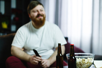 Happy fat man sits on the sofa and watches TV with popcorn and beer