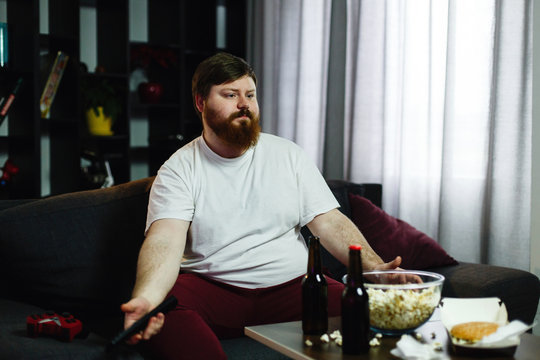 Happy fat man sits on the sofa and watches TV with popcorn and beer