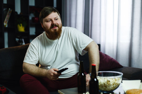 Happy fat man sits on the sofa and watches TV with popcorn and beer