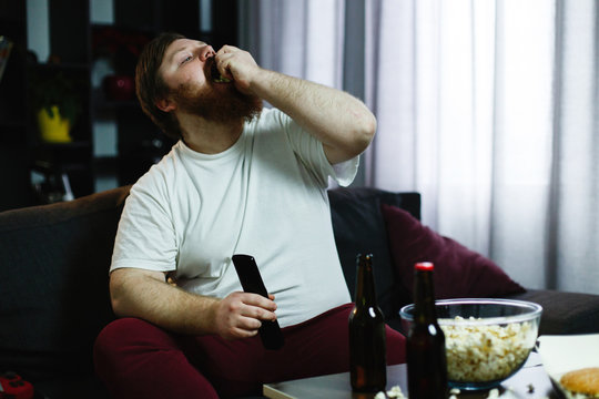 Happy fat man eats pop-corn lying on the sofa before a table with beer