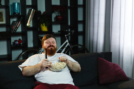 Laughing fat man sits on the sofa, eats pop-corn and watches TV
