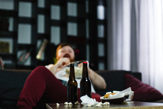 Happy fat man eats pop-corn lying on the sofa before a table with beer