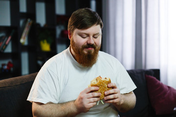 Smiling fat man eats burger sitting before a TV-set