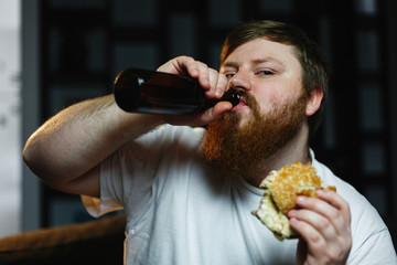 Fat man watches TV, eats burger and drinks beer