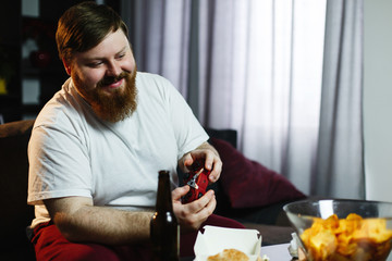 Obraz na płótnie Canvas Happy fat man in dirty shirt plays video games drinking beer and eating pop-corn and chips