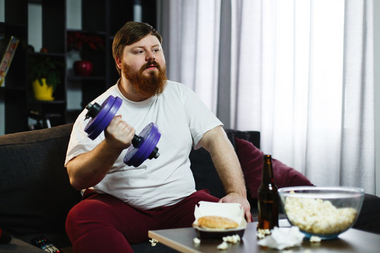 Fat man doing sport while he sits with food before a TV-set
