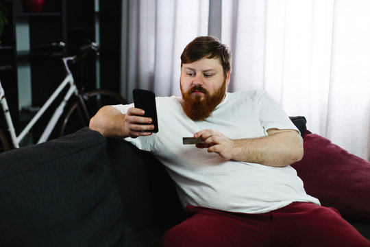 Fat man types the number of a credit card in his phone sitting on the couch
