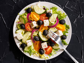 Plate Greek salad with a fork on the table, top view. Fresh diet vegetarian salad decorated with herbs on a dark stone slate background.