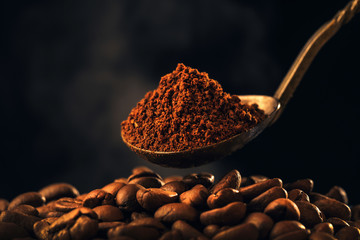 Roasted ground coffee into an old silver spoon and roasting coffee beans with smoke on dark background. Close-up photo. Vintage style effect picture, with copy space.