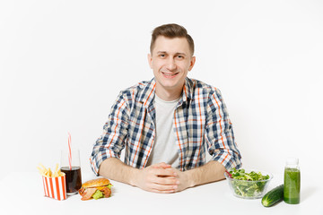Man sitting at table green detox smoothies, salad in glass bowl, cucumber, burger, fries, cola in glass isolated on white background. Proper nutrition, healthy lifestyle, fast food, choice concept.