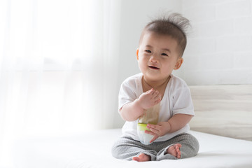 Asian baby boy holding bottle milk and sitting on white bed