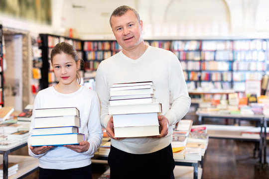 Portrait of tweenage girl with father standing in library with pile of books in hands