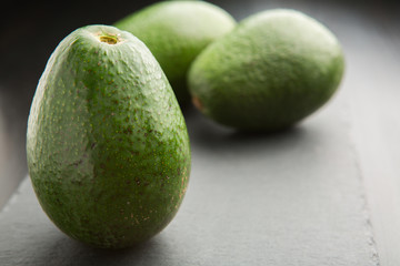 Three raw fresh avocados in black ceramic plate on black concrete background. Selective focus.
