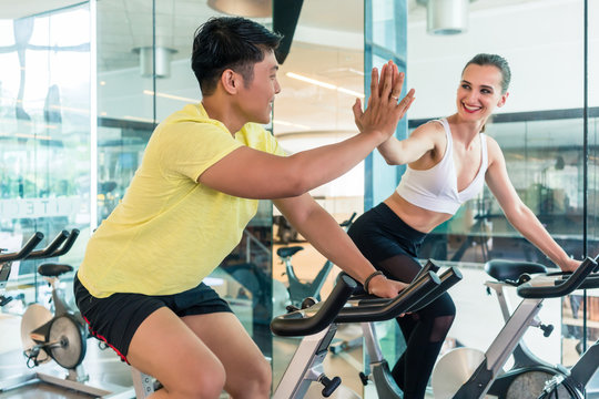 Cheerful young man and his beautiful workout partner and friend giving high five during indoor cycling workout in a trendy fitness club