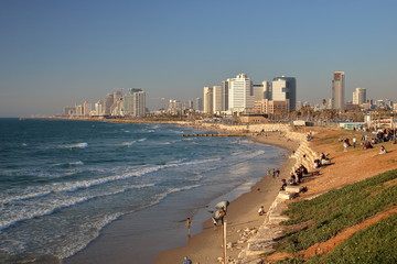 Widok na zatokę Morza Śródziemnego, plażę, nabrzeże ze spacerującymi i odpoczywającymi ludźmi, w tle wysokie nowoczesne budynki, Tel Awiw, Izrael, fale na morzu, błękitne niebo