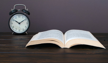 Still life photo of  an open book with black alram clock on wooden table