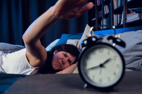 Picture of dissatisfied woman with insomnia stretching arm to alarm clock at night