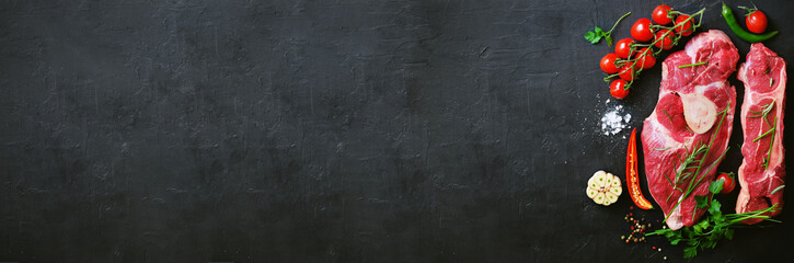 Raw meat steaks with cherry tomatoes, hot pepper, garlic, oil and herbs on dark stone, concrete background. Banner