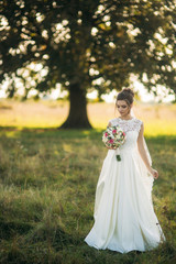 Fototapeta na wymiar Beautiful bride with bouquet standing in the middle of field in the background of big tree