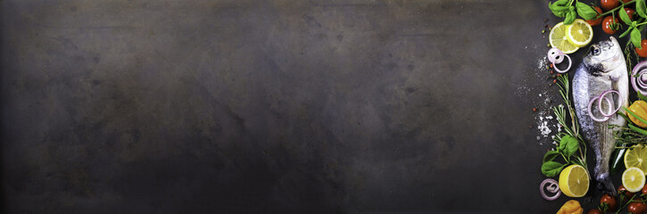 Fresh uncooked fish, dorado, sea bream with lemon, herbs, vegetables and spices on stone background. Top view. Banner
