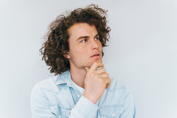 Studio shot of handsome pensive male model with curly hair, wears blue shirt, looking up thinking about problem, holding his chin on white. People, business concept. Copy space for your advertisement
