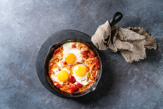 Traditional Israeli Cuisine dishes Shakshuka. Fried egg with vegetables tomatoes and paprika in cast-iron pan on wooden board with cloth over blue texture background. Top view, space.