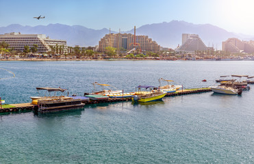 Naklejka premium Eilat at summer day. View on hotels zone and sand beaches in the back and water sport floating pier with motor-boats for rent in the front.