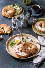 Variety of homemade bagels with sesame seeds, cream cheese, pesto sauce, eggs, radish, herbs served on ceramic plate with ingredients and coffee above over blue texture background. Top view, space.