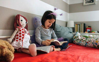 Little girl disguised as a butterfly sitting on the bed reading a book to her rag doll