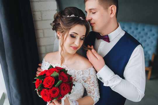 Beautiful newlyweds in the studio near the window. The groom tenderly embraces the bride in the gray studio and straightens her hair. Portrait of a newlywed couple.