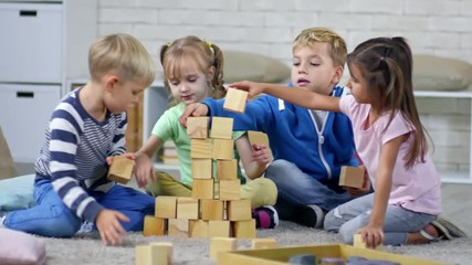 Group of four playful primary school children sitting together on the floor and building tower with wooden toy blocks - Powered by Adobe
