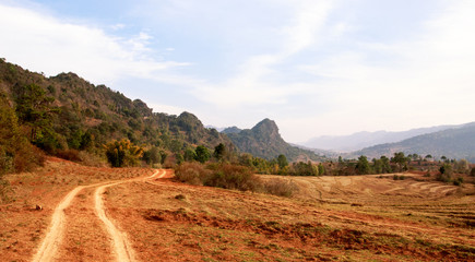 Fototapeta premium Dirt road winds between arid farmland and low mountain range in Shan State, Myanmar
