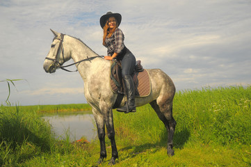 girl in plaid shirt and cowboy hat riding a horse by the lake