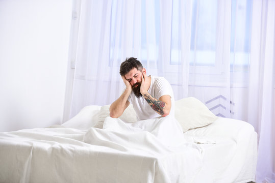 Man sitting on bed, falling asleep while lean on hand, white curtains on background. Handsome tired man leaning on his hand and sitting with closed eyes on his bed while getting up in the morning.