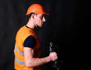 Repairman holds dumbbell in his right hand, black background. Strong builder concept. Builder in helmet working out, copy space. Worker, contractor, builder on strict face with muscular biceps.