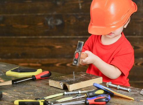 Kid boy hammering nail into wooden board. Toddler on busy face plays at home in workshop. Handcrafting concept. Child in too big helmet cute playing as builder or repairer, repairing or handcrafting.