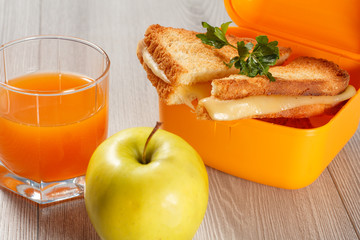 Yellow sandwich box with toasted slices of bread, cheese and green parsley, green apple and glass of orange juice on wooden desk