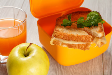 Yellow sandwich box with toasted slices of bread, cheese and green parsley, green apple and glass of orange juice on wooden desk
