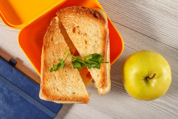 Yellow sandwich box with toasted slices of bread, cheese and green parsley, green apple and hardback books on the background
