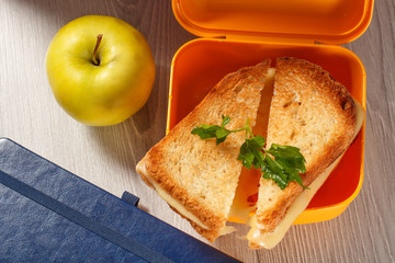 Yellow sandwich box with toasted slices of bread, cheese and green parsley, green apple and hardback books on the background