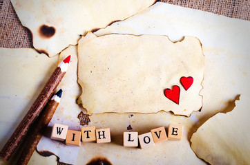 Old vintage sheet of paper, two red hearts, wooden pencils and words With love on cubes on burlap, sackcloth background. Retro design effects.