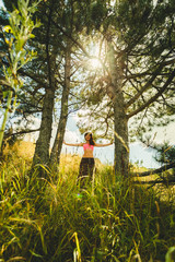 A young beautiful woman with an ideal figure enjoys the freshness of a summer day in a pine grove among dense grass. The sun is sparkling in the midst of the trees.
