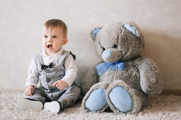 Cute happy baby in stylish gentleman costume sits on a white carpet and plays with her Teddy bear, laughing at the baby.