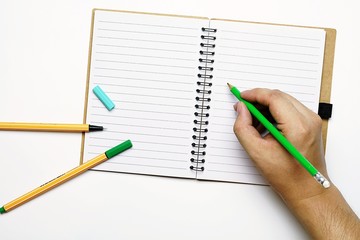top view image of pens, pencil and book with copy space, A hand holding a green pencil, trying to write something on a book, that was opened on white table.