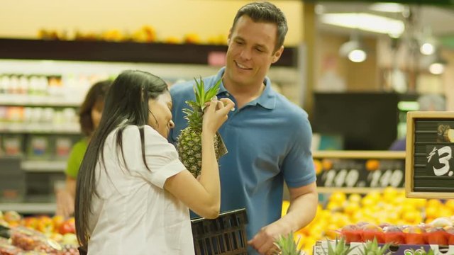 A couple smell a pineapple together as they pick out produce in a grocery store