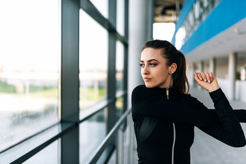 Concentrated slim woman in sports clothing stretching her arms while standing near the window.