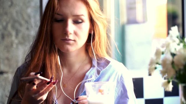 Pretty woman with red hair listening music on smartphone in cafe, super slow motion
