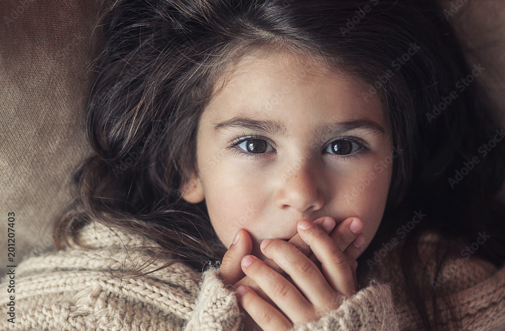 Wall mural little beautiful child girl with big brown eyes lies and closes her mouth with her hands in silence