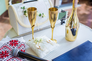 Two gold wedding glasses for champagne, golden bottle of champagne. Two gold wedding rings on pillow.