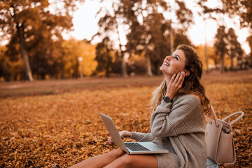Gorgeous young woman listening music while sitting with laptop in the autumn park.